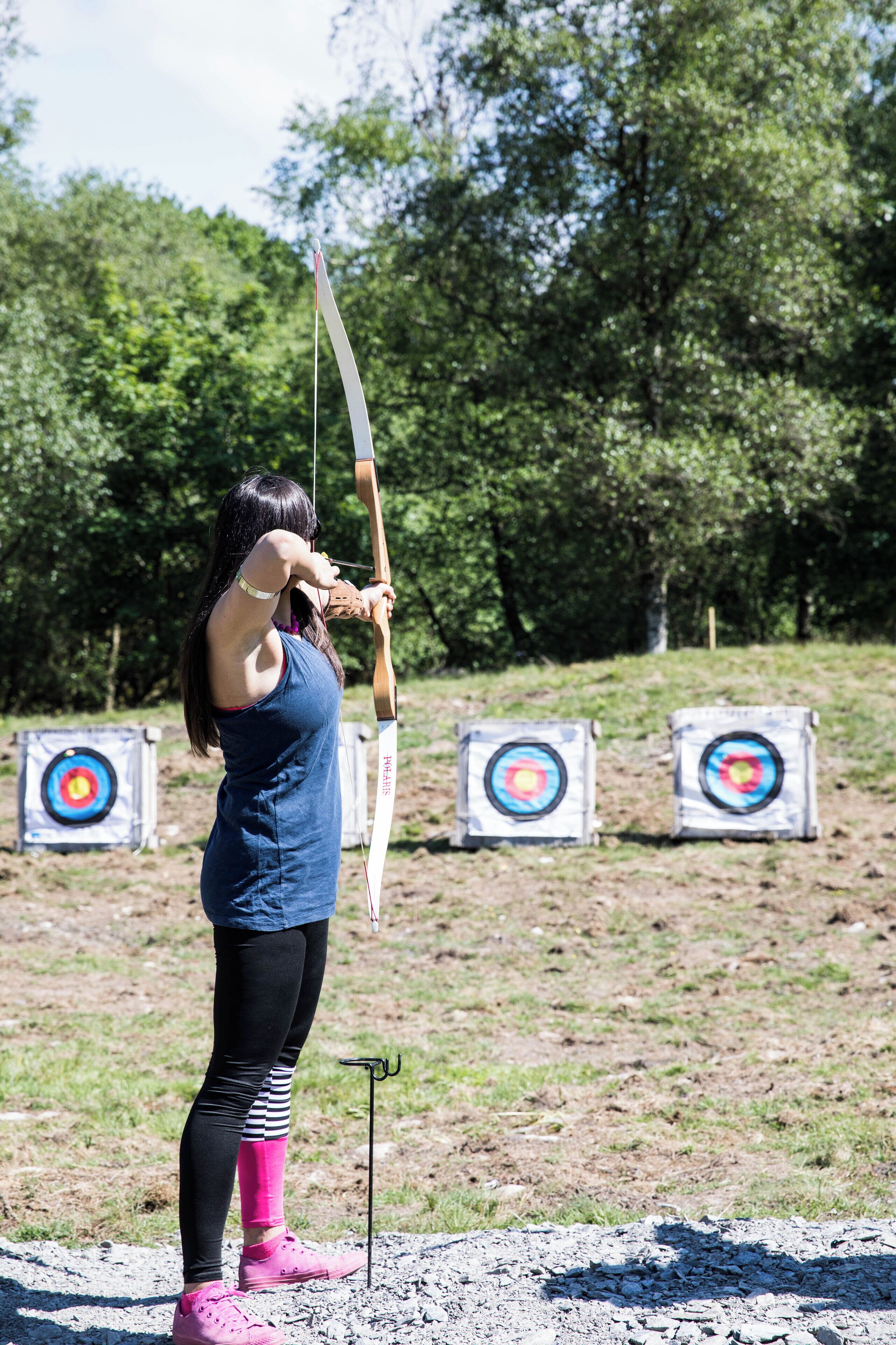 A person in a blue tank top and black leggings with pink shoes, aiming a bow at a target.