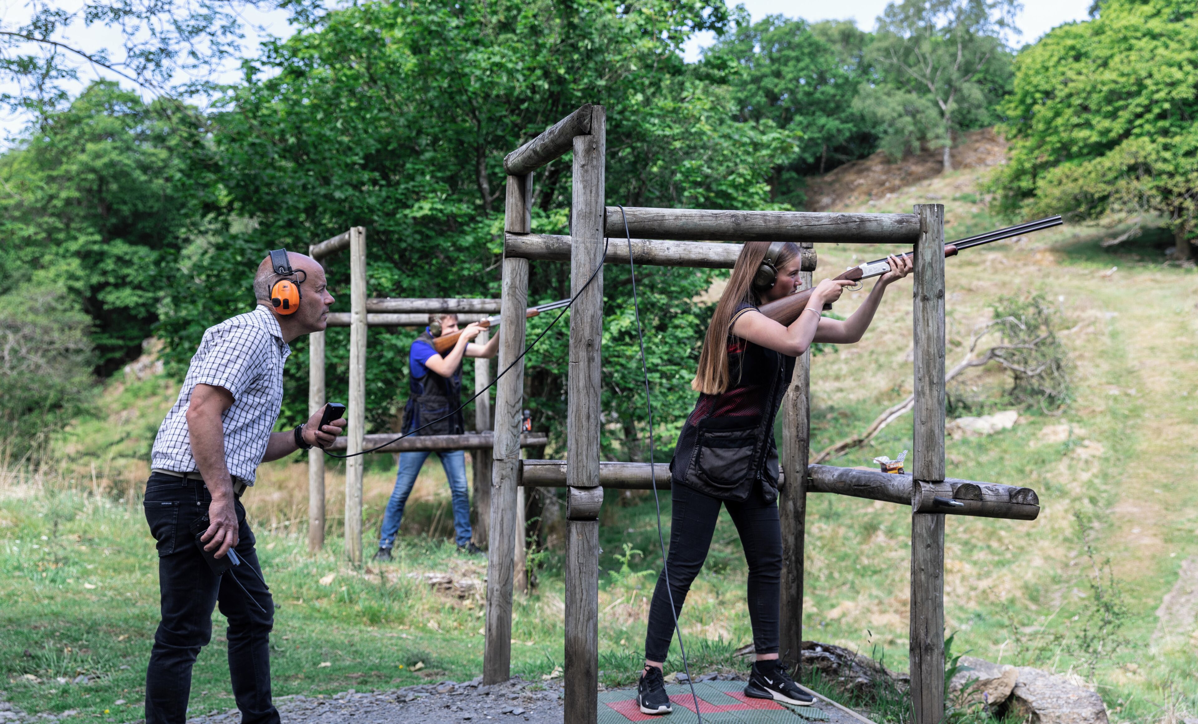 A person aiming a shotgun at a wooden target structure outdoors.