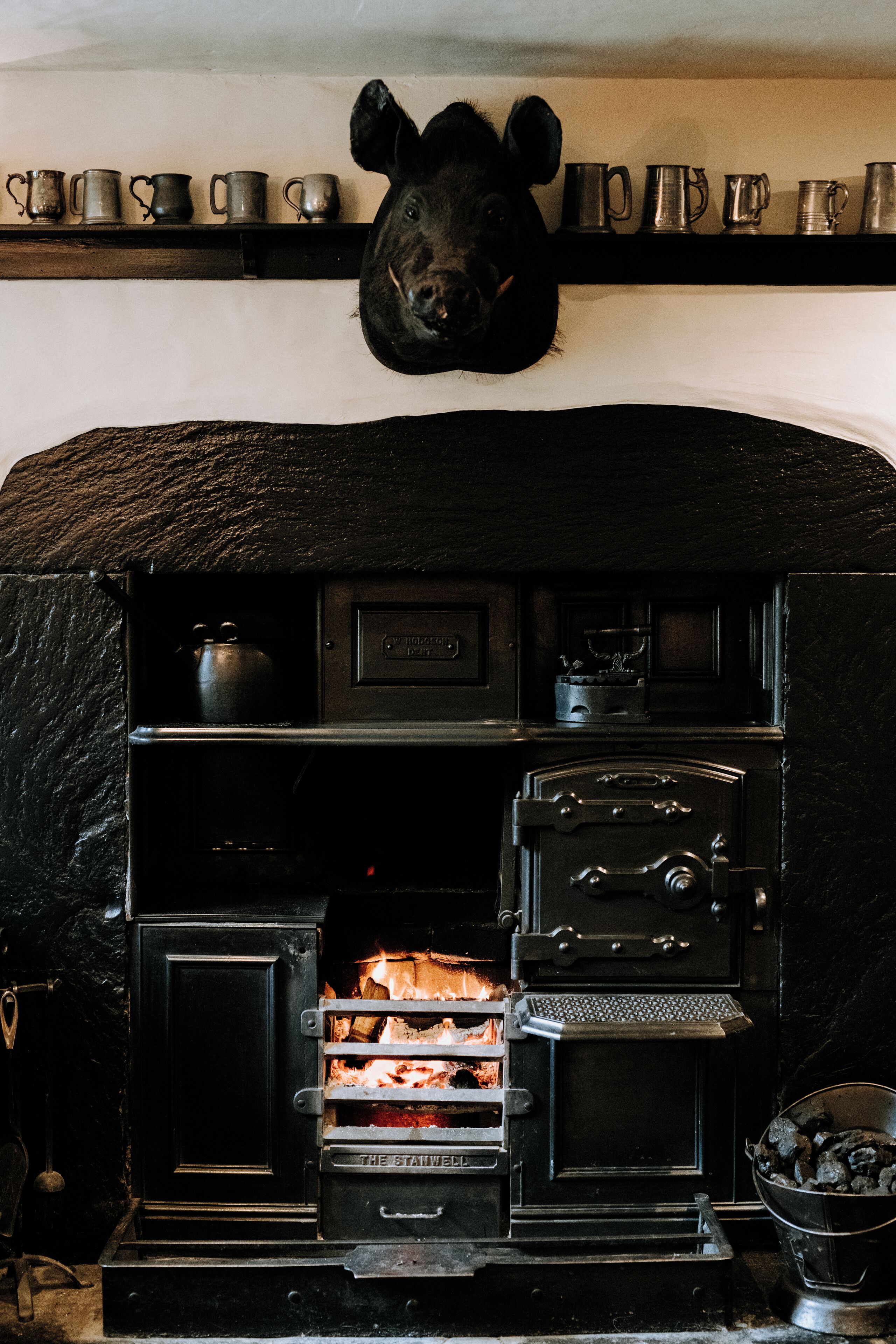 A traditional fireplace with a black cow head decoration above it, a shelf with metal mugs, and a wood-burning stove below.