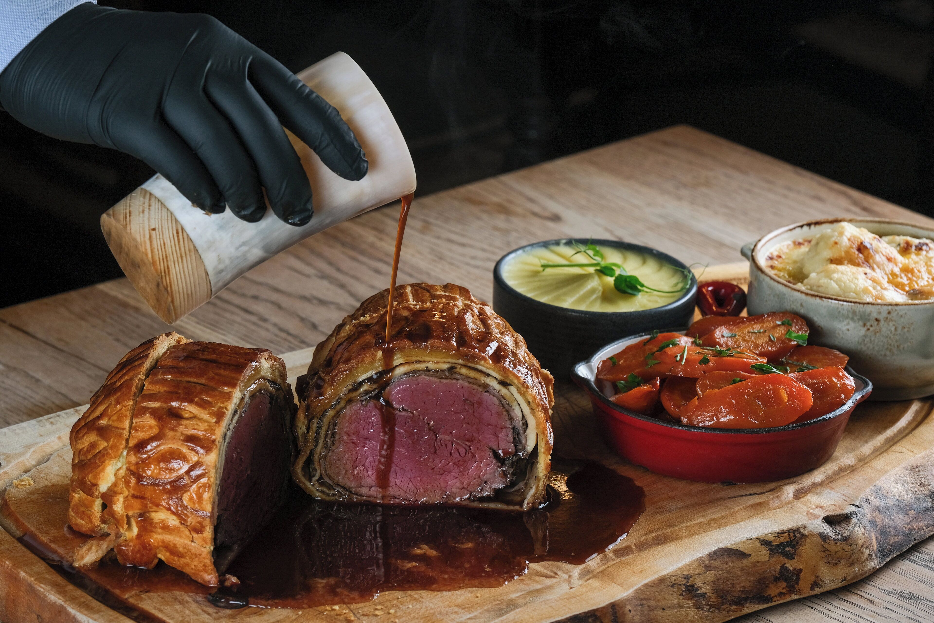 A chef pouring sauce over a roast beef dish, accompanied by vegetables and a bowl of mashed potatoes.