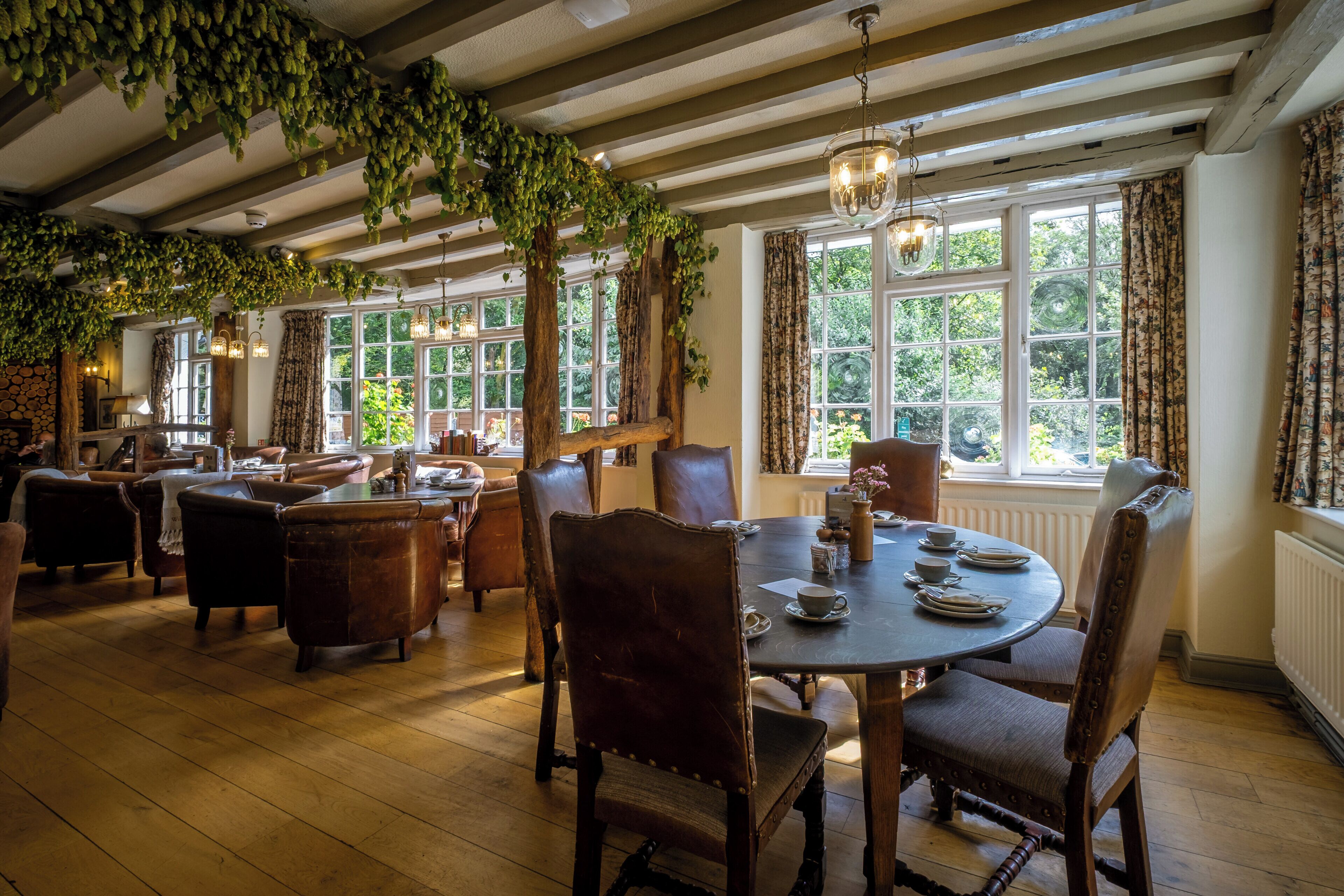 A dining area with wooden tables and chairs, large windows with curtains, and hanging greenery.