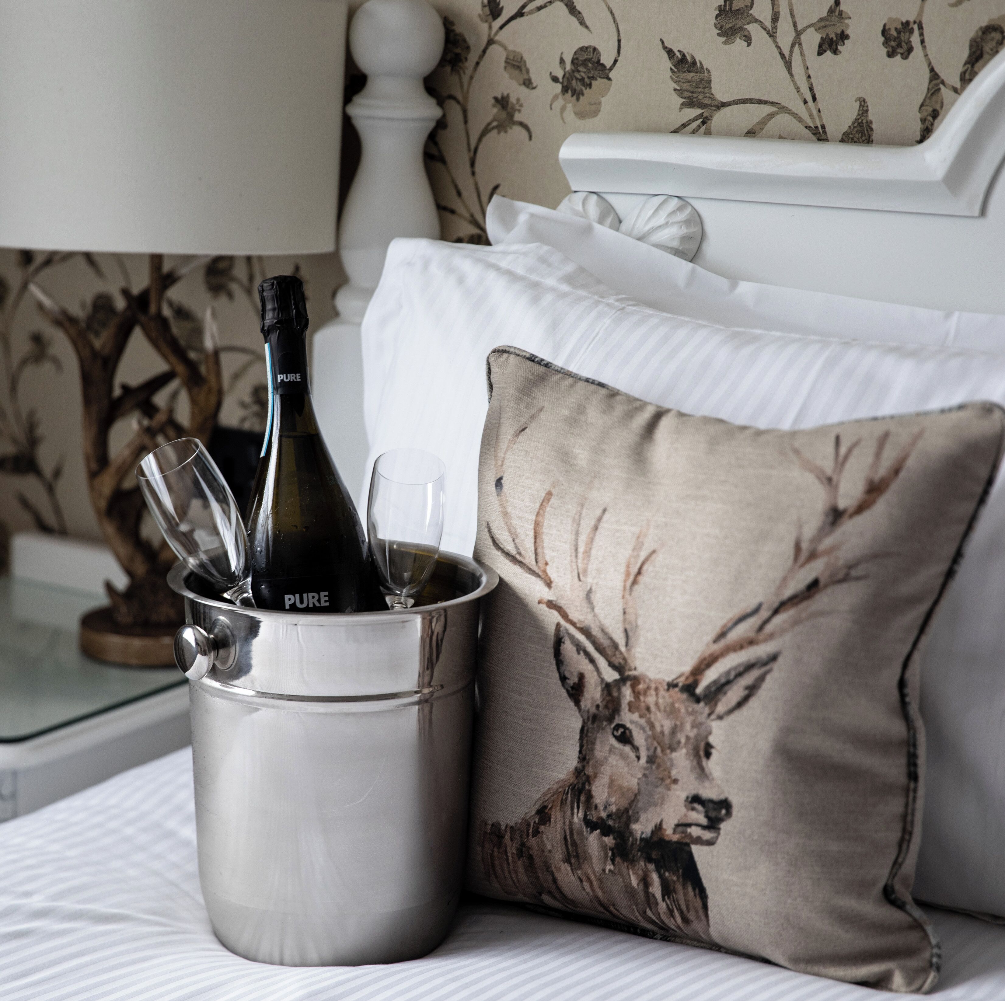 Een bed met een metalen emmer, daarin een fles wijn en een glas, een kussen met een hertenprint, en een behang met bloemen.