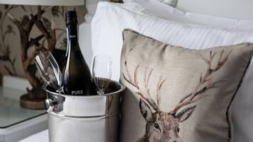 Een bed met een metalen emmer, daarin een fles wijn en een glas, een kussen met een hertenprint, en een behang met bloemen.
