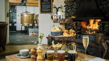 A wooden table with a variety of food items, drinks, and a fireplace in the background.