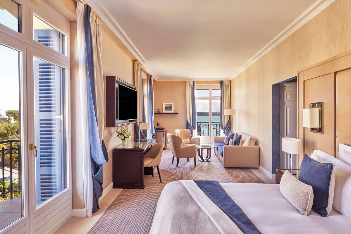 A hotel room with a large bed, a desk with a TV, a sofa, and a balcony with curtains.