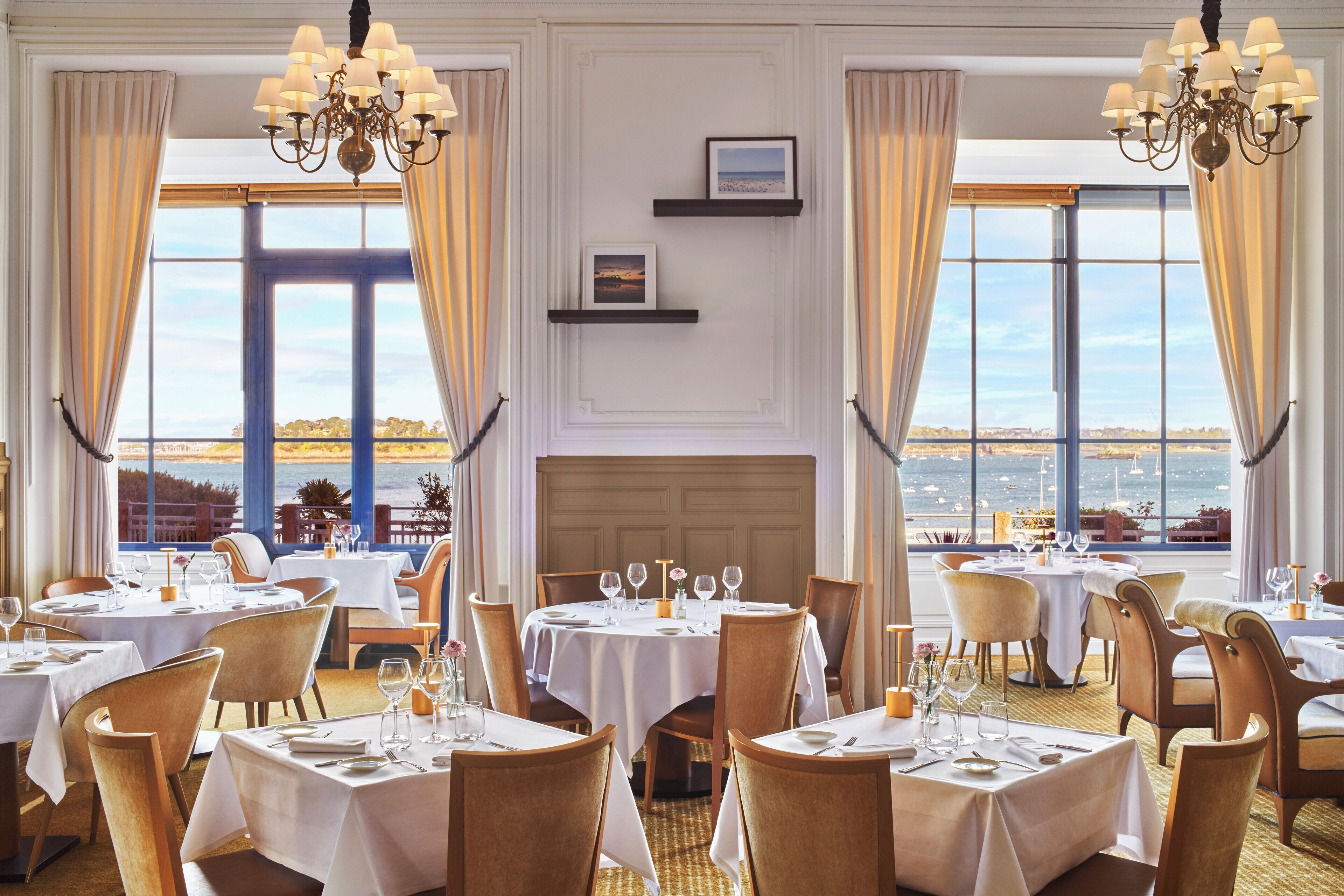 Un espace repas avec des tables dressées pour le repas, de grandes fenêtres avec des rideaux et une vue sur la mer.