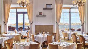 Un espace repas avec des tables dressées pour le repas, de grandes fenêtres avec des rideaux et une vue sur la mer.