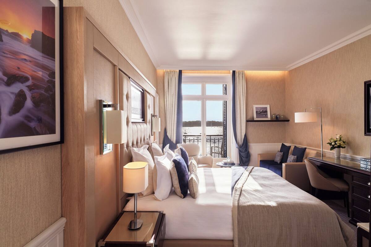 A hotel room with a large bed, a desk, a chair, a lamp, and a view of the outside.