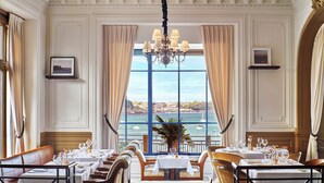 Comedor con mesas dispuestas para una comida, una araña y vista al mar a través de amplios ventanales.