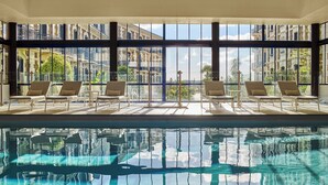 Una piscina interior con paredes de vidrio transparente, sillones de descanso y vistas de edificios circundantes.