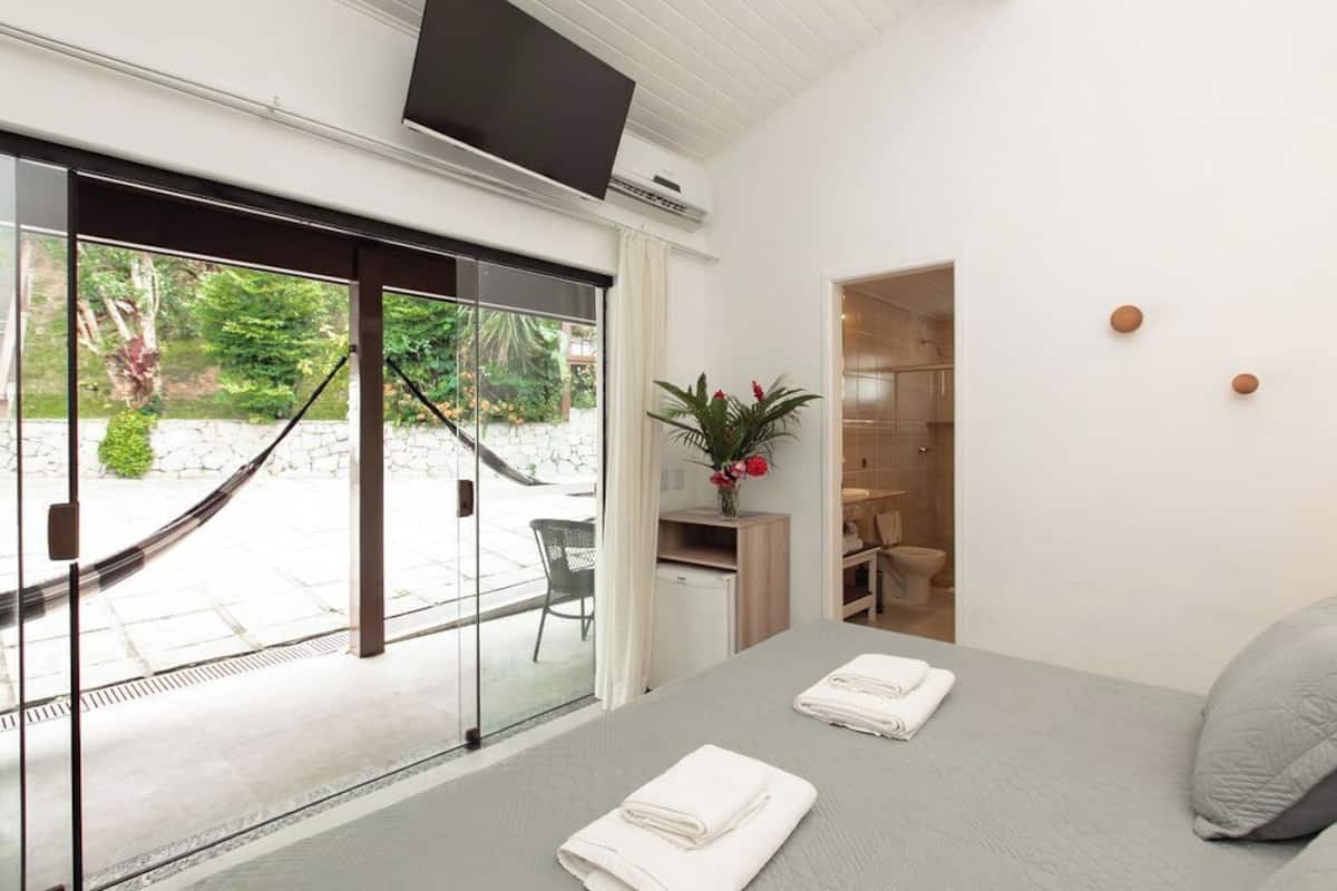 Ett modernt hotellrum med en stor skjutdörr i glas som leder ut till en uteplats, en säng med prydligt vikta handdukar, ett litet skrivbord med en växt och en tv som är monterad på väggen.