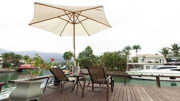 Een houten terras met een tafel en stoelen, een grote parasol, en uitzicht op een jachthaven met boten en palmbomen.