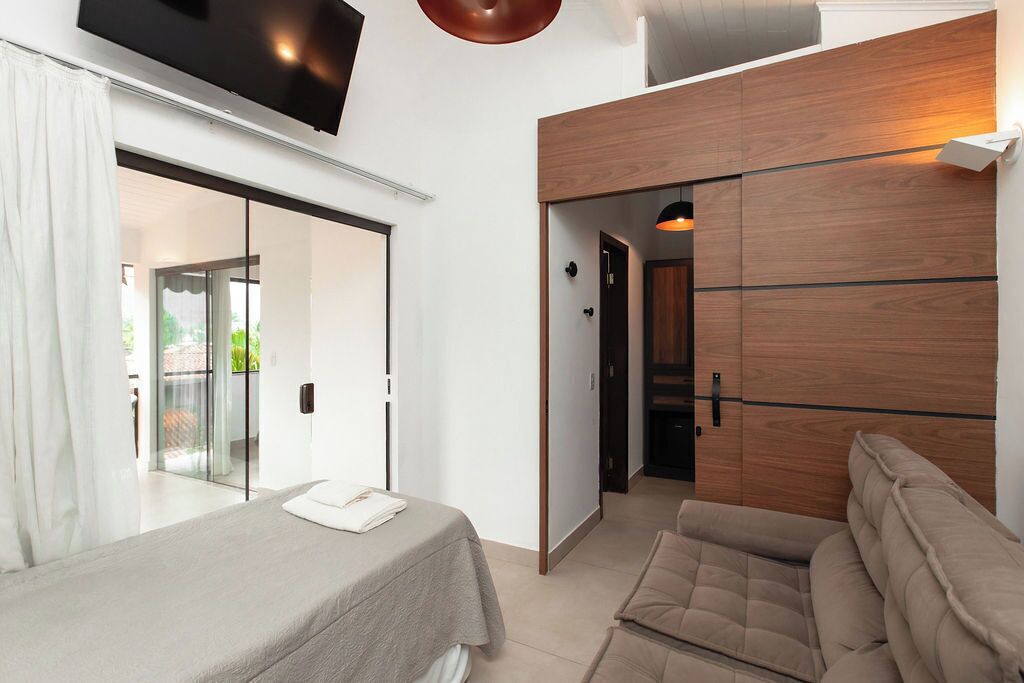 Quarto moderno com cama, sofá, televisão e porta de correr para uma varanda.