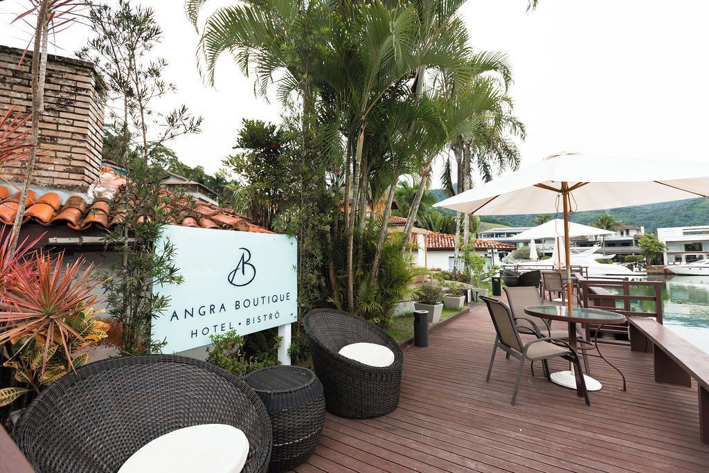 A wooden deck with wicker furniture, a sign for Angra Boutique Hotel & Bistro, and a view of boats in the background.