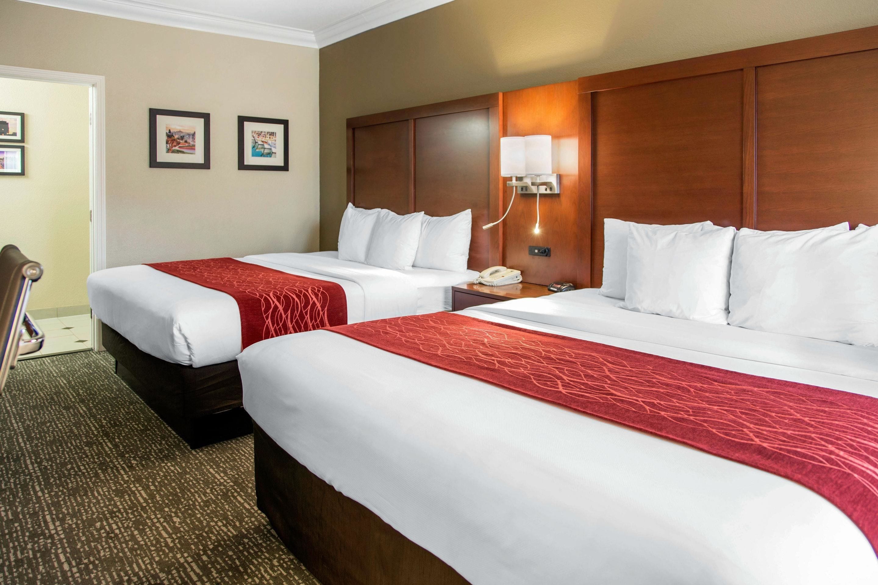 A hotel room with two beds, a wooden headboard, a bedside table with a lamp, and framed pictures on the wall.