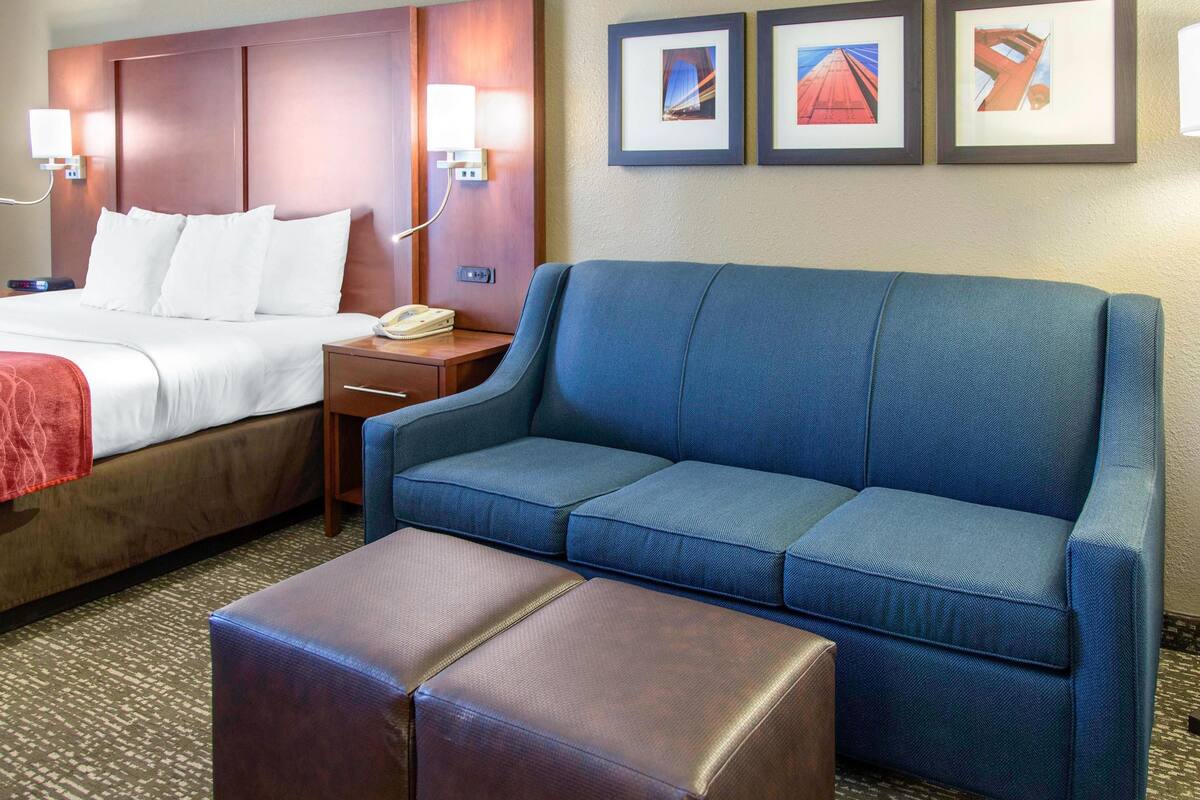 A hotel room with a bed, a blue sofa, a brown ottoman, and a nightstand with a phone.