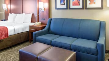 A hotel room with a bed, a blue sofa, a brown ottoman, and a nightstand with a phone.