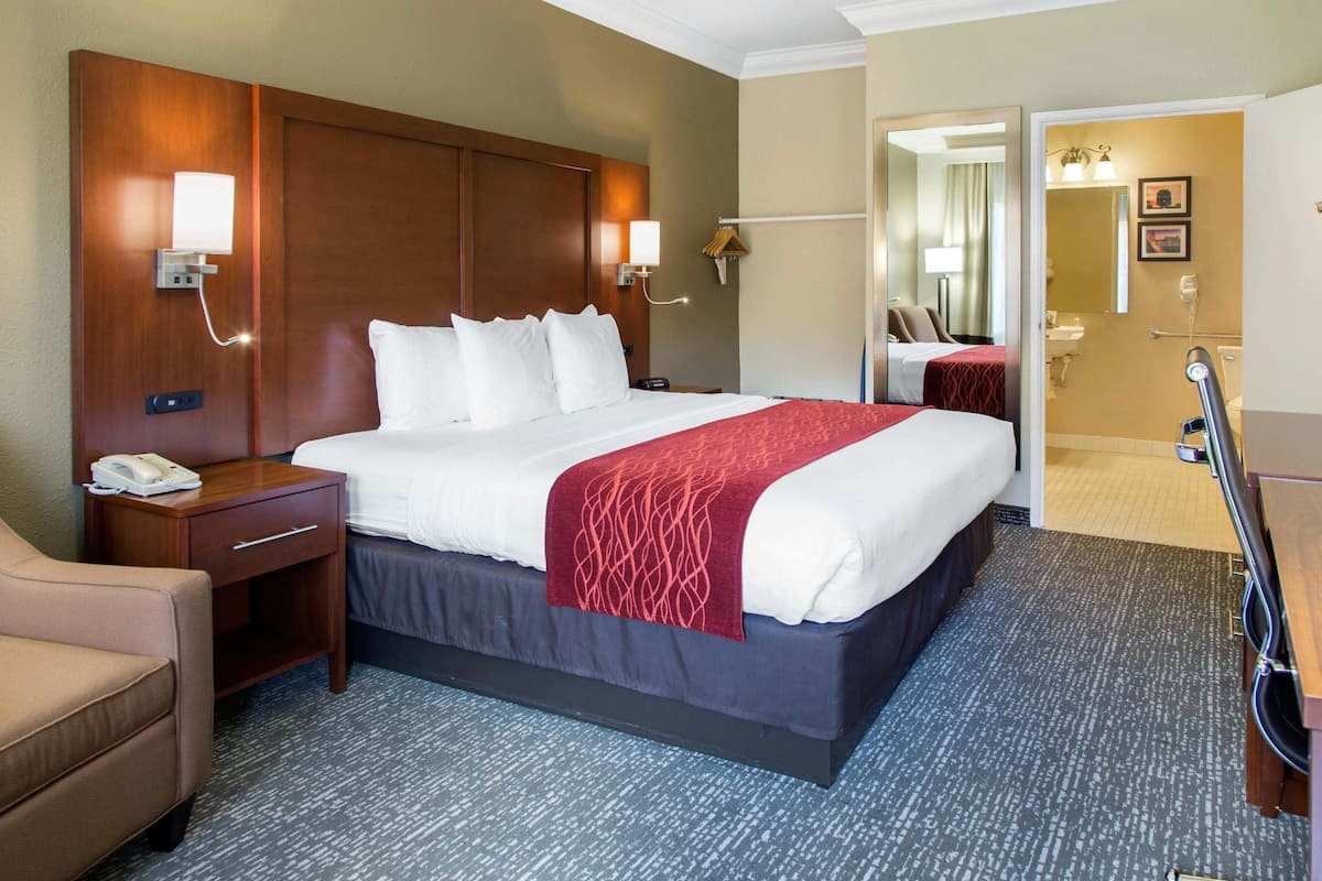 A hotel room with a large bed, a bedside table with a phone, a desk lamp, a chair, and a bathroom visible through an open door.