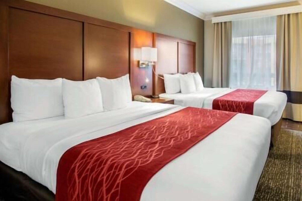 Habitación de hotel con dos camas, cabecero de madera, mesita de noche con lámpara y ventana con cortinas.