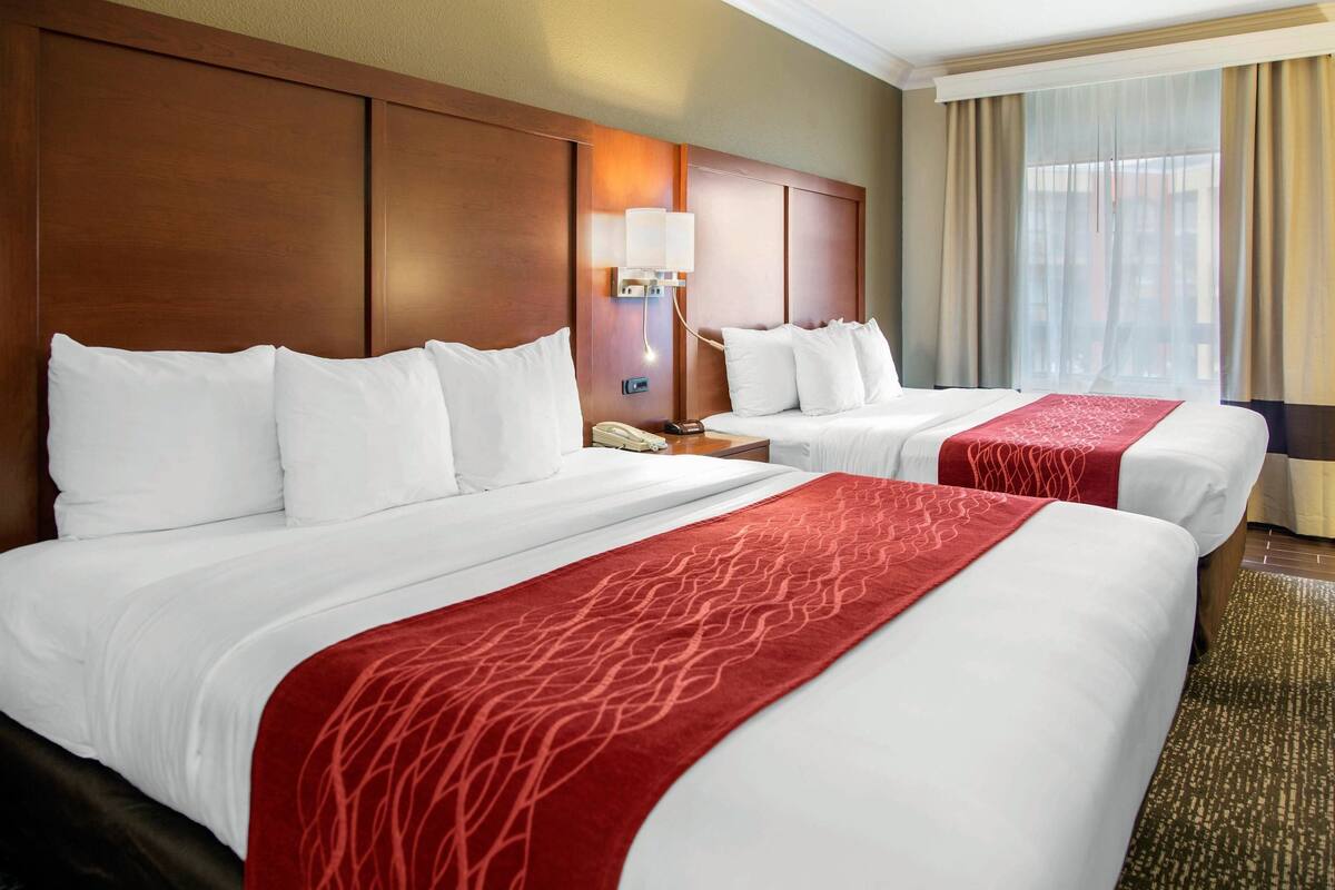 A hotel room with two beds, a wooden headboard, a bedside table with a lamp, and a window with curtains.