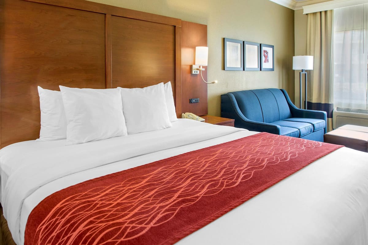 A hotel room with a large bed, a blue armchair, a bedside table with a lamp, and a wall-mounted phone.