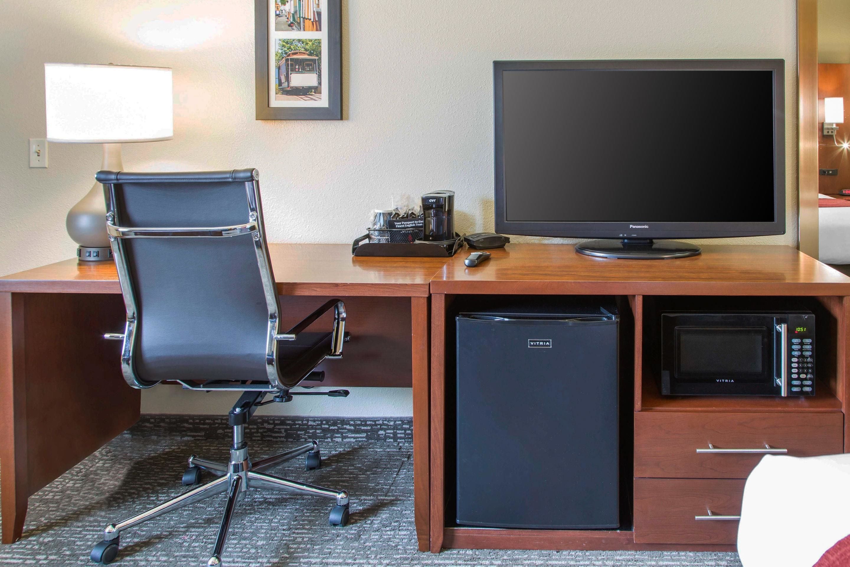 Quarto de hotel com escrivaninha de madeira, cadeira de escritório preta, televisão de tela plana, micro-ondas e mini-geladeira.