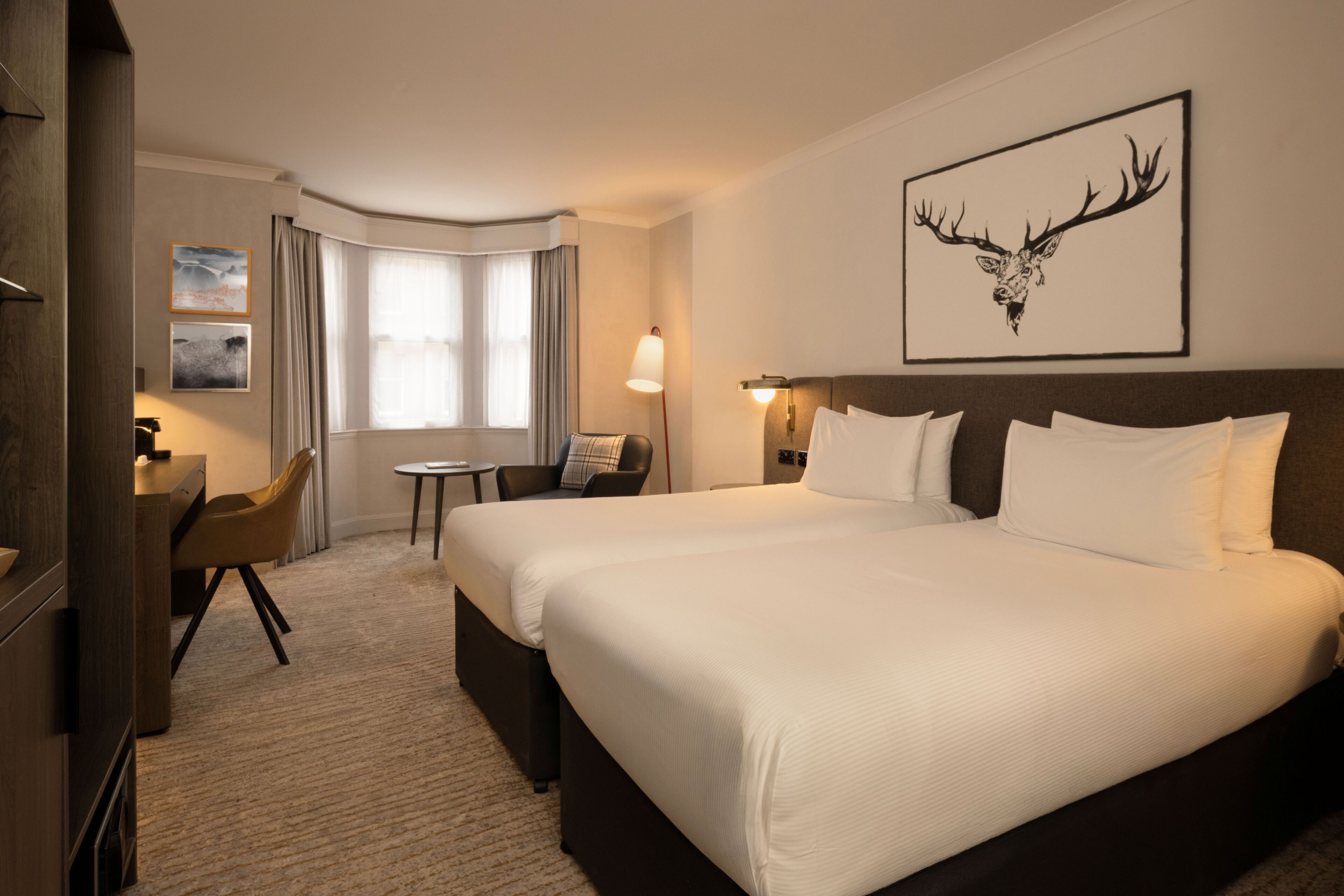 A hotel room with a large bed, a desk, a chair, a small table, and a window with curtains.