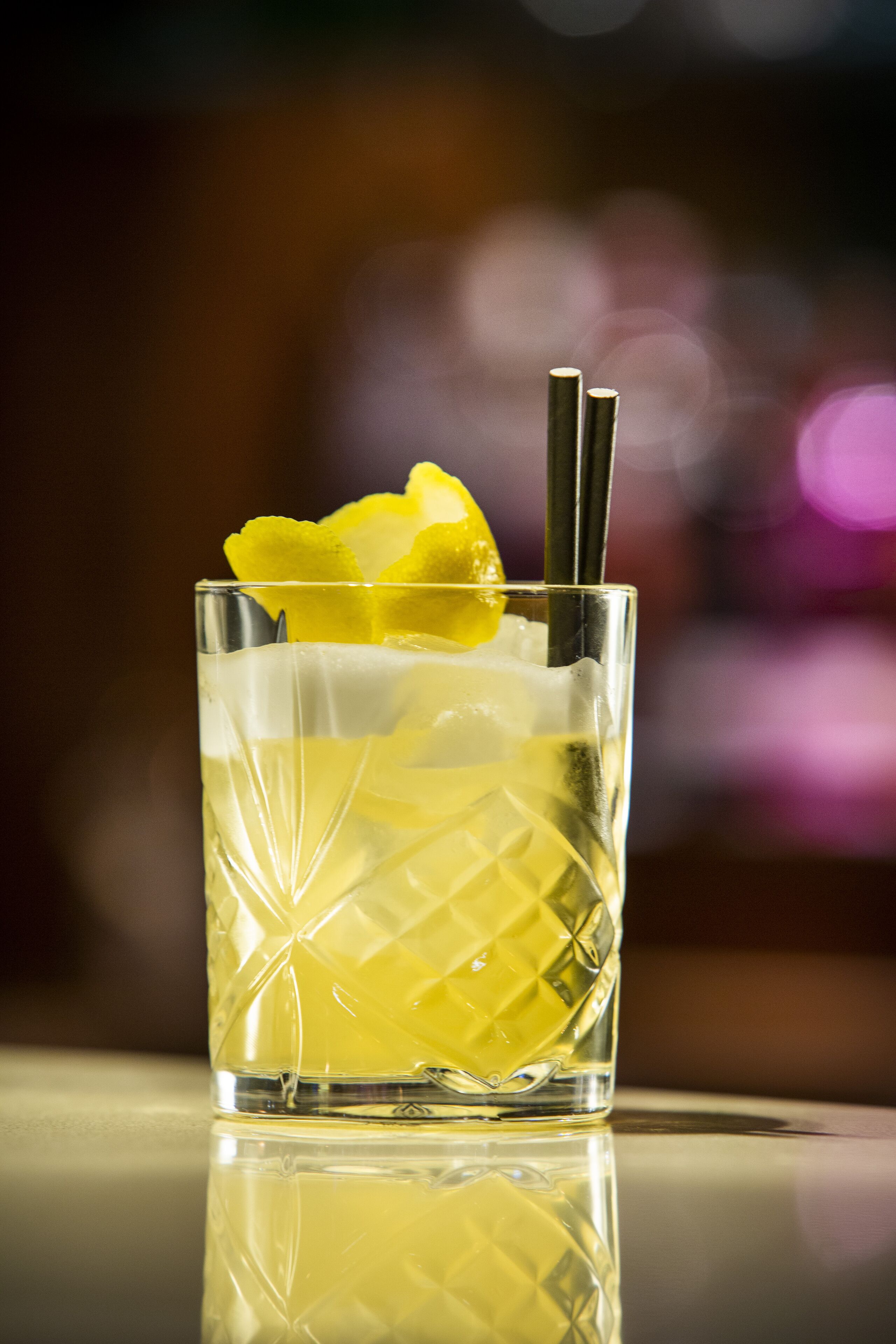 A clear glass with a textured pattern, containing a yellow beverage, a lemon slice, and two straws.
