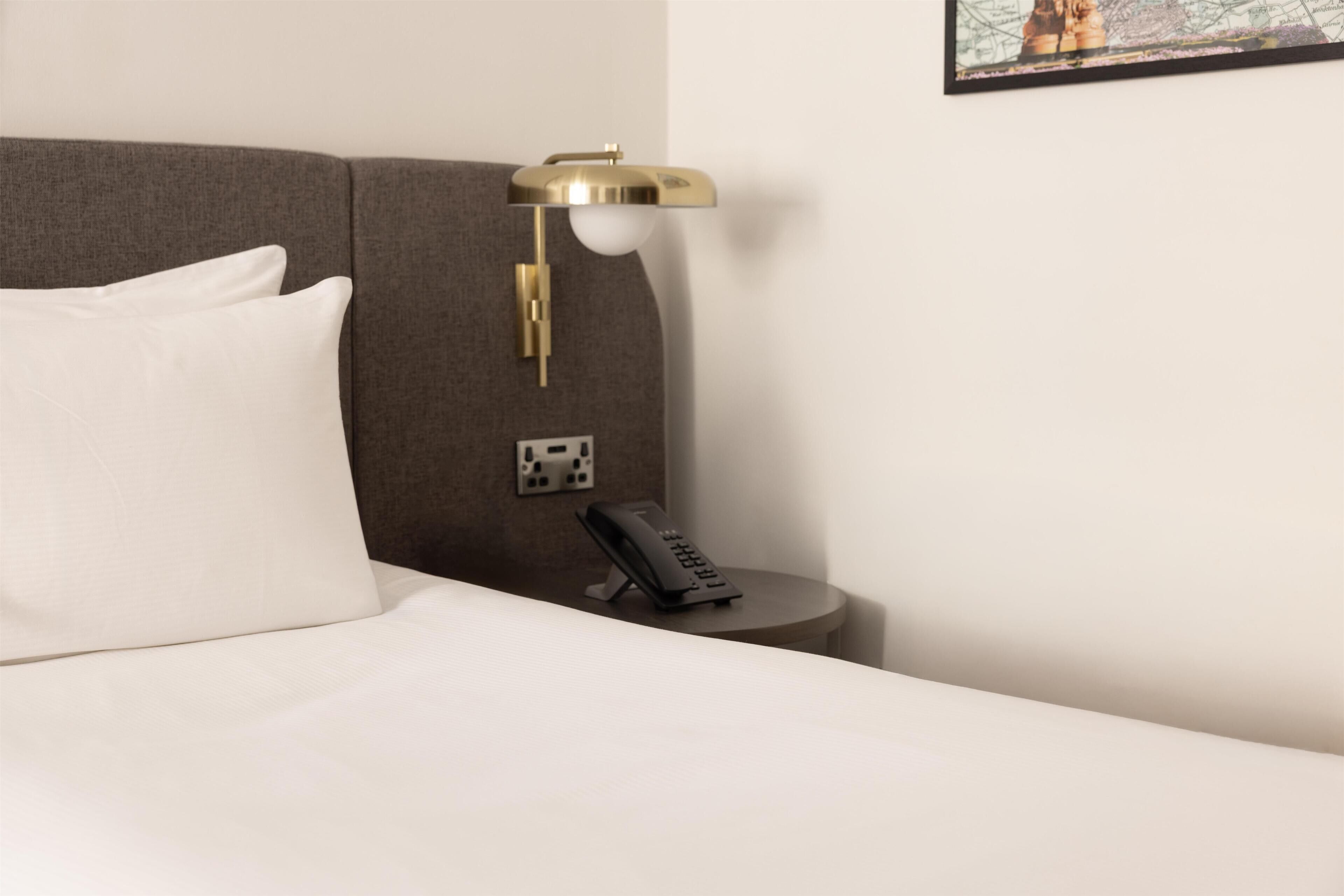 A neatly made bed with white bedding, a brown headboard, a small bedside table with a black telephone, and a gold-colored wall-mounted lamp.