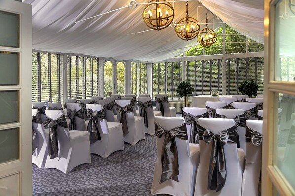 Un espacio para eventos con carpas, filas de sillas blancas decoradas con fajines negros, ventanas amplias y candelabros colgantes.