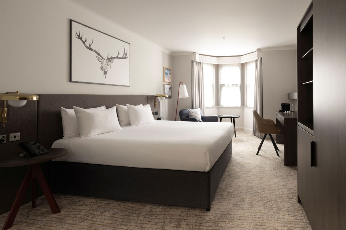 Une chambre d’hôtel avec un grand lit, un bureau, une chaise, une lampe et une fenêtre avec des rideaux.