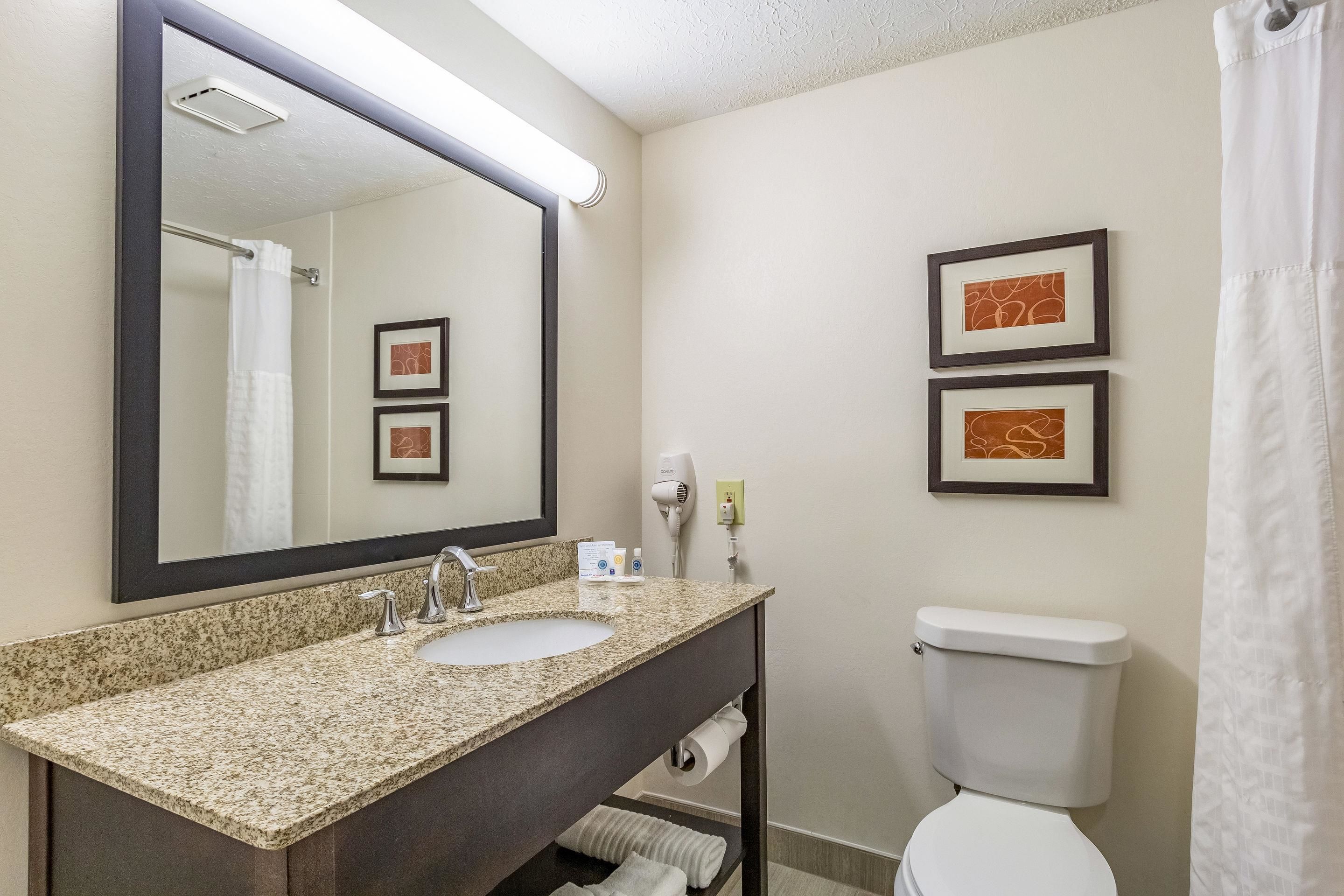A bathroom with a granite countertop sink, a mirror, a toilet, and two framed pictures on the wall.