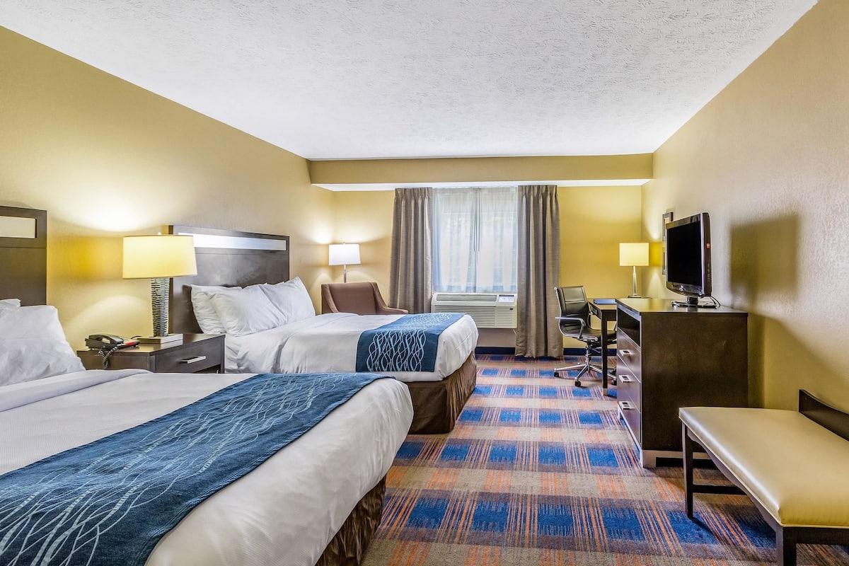 A hotel room with two beds, a desk, a TV, and a bench.