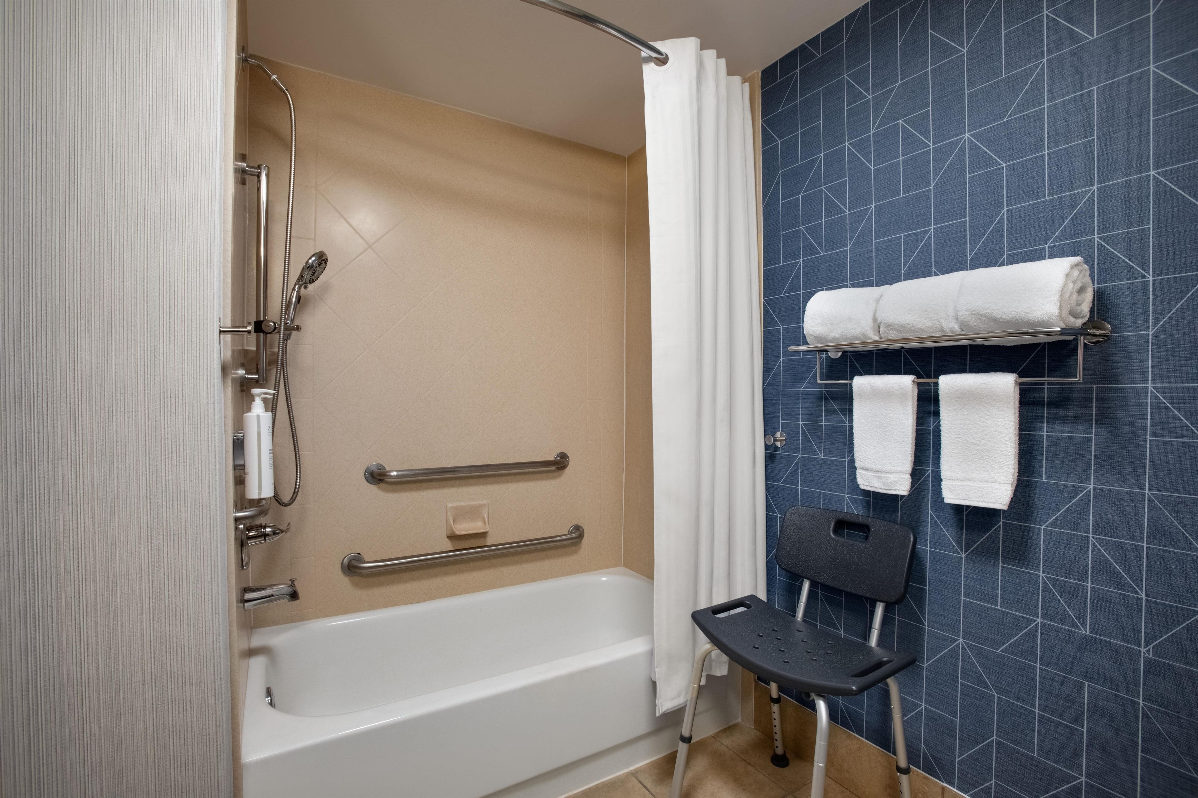 A bathroom with a bathtub, a shower, a handrail, a towel rack, and a black chair.
