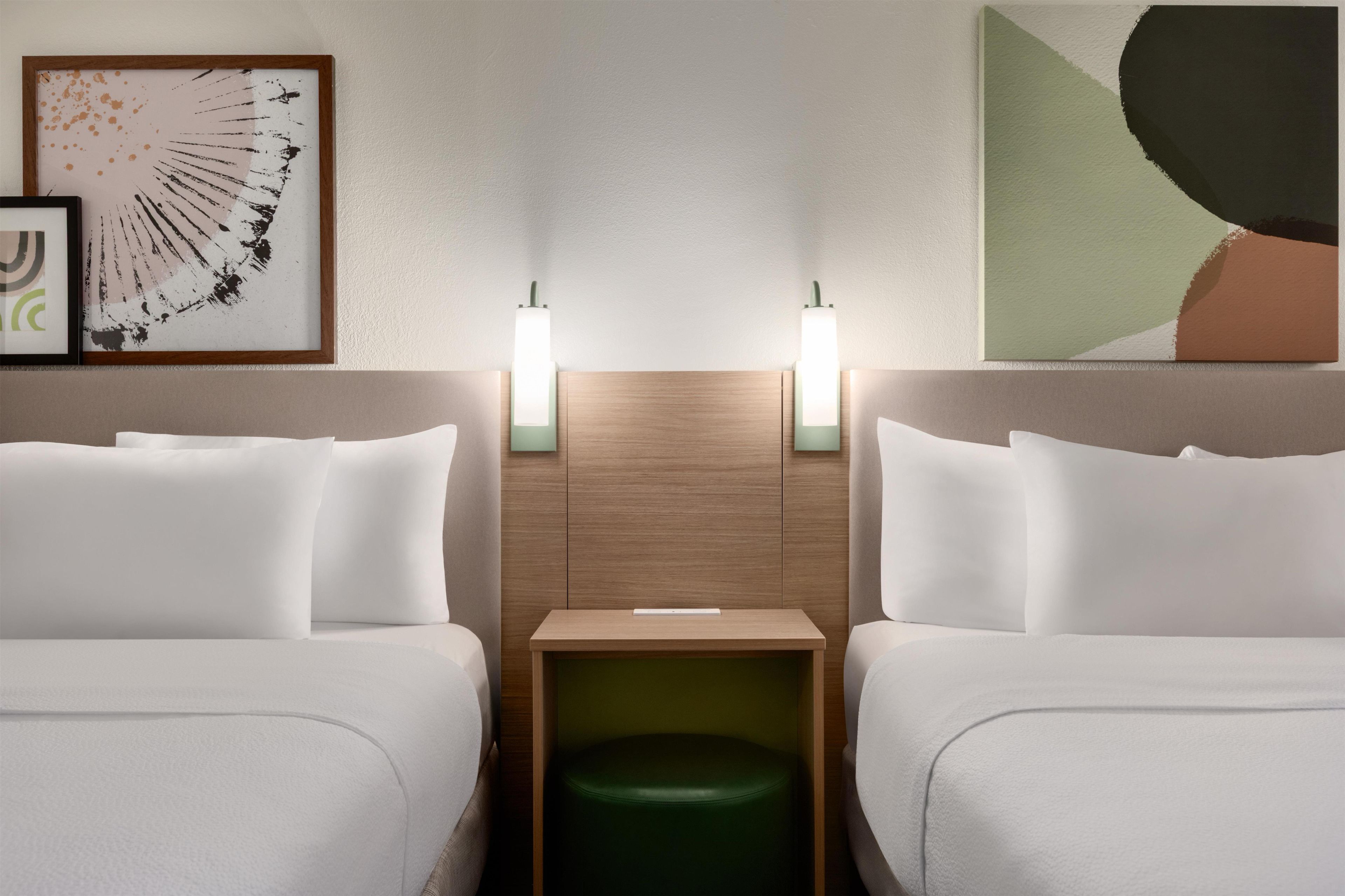 A hotel room with two beds, a nightstand, a green ottoman, and two wall-mounted lights.