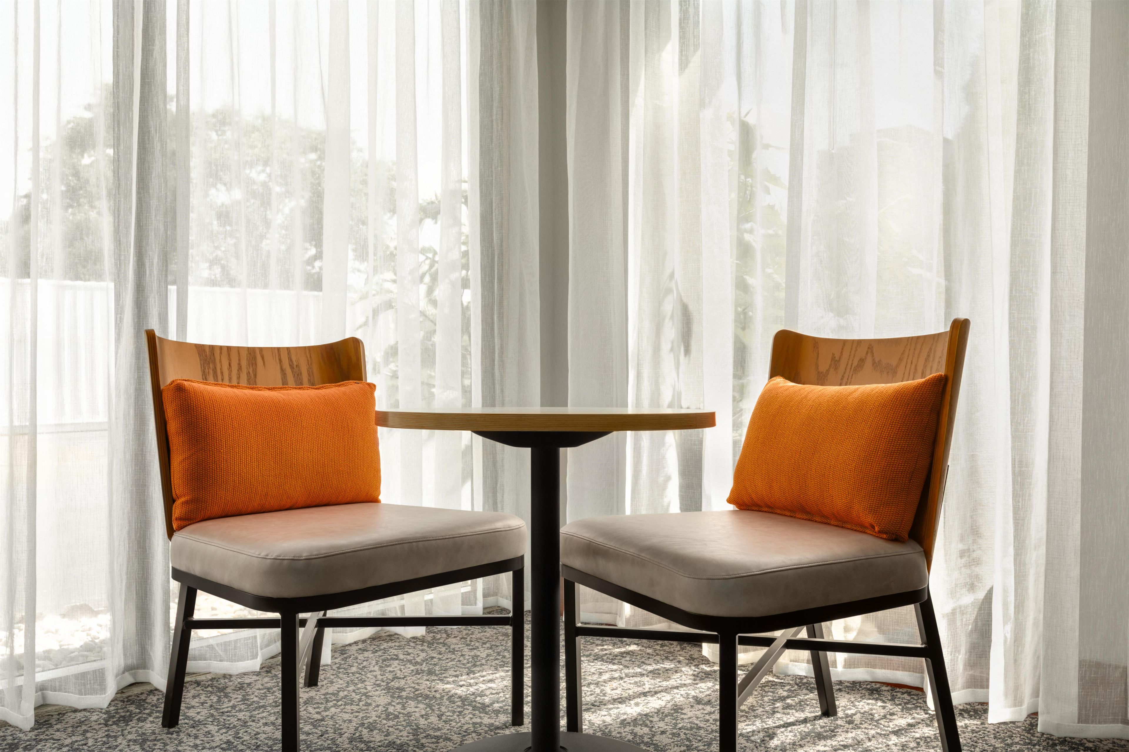 Two chairs with orange cushions and a small round table in front of a window with sheer curtains.