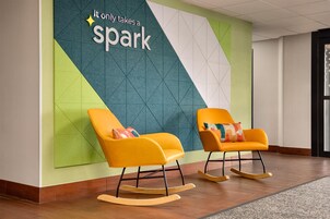 Um lobby moderno com duas poltronas de balanço, design geométrico na parede e uma placa com a mensagem 'Tudo o que é preciso é uma faísca'.