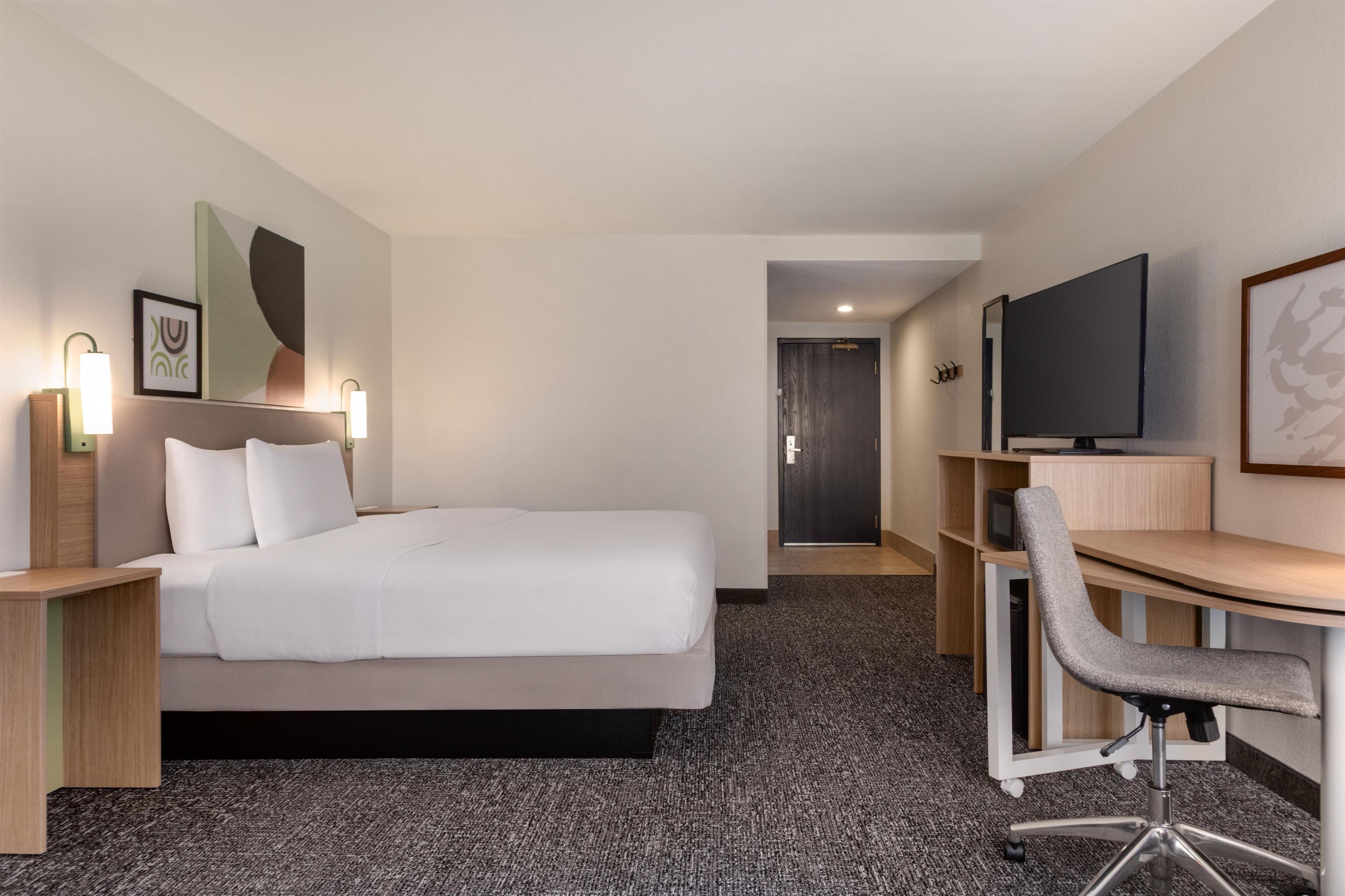 A hotel room with a bed, bedside tables, a desk with a chair, a television, and artwork on the walls.