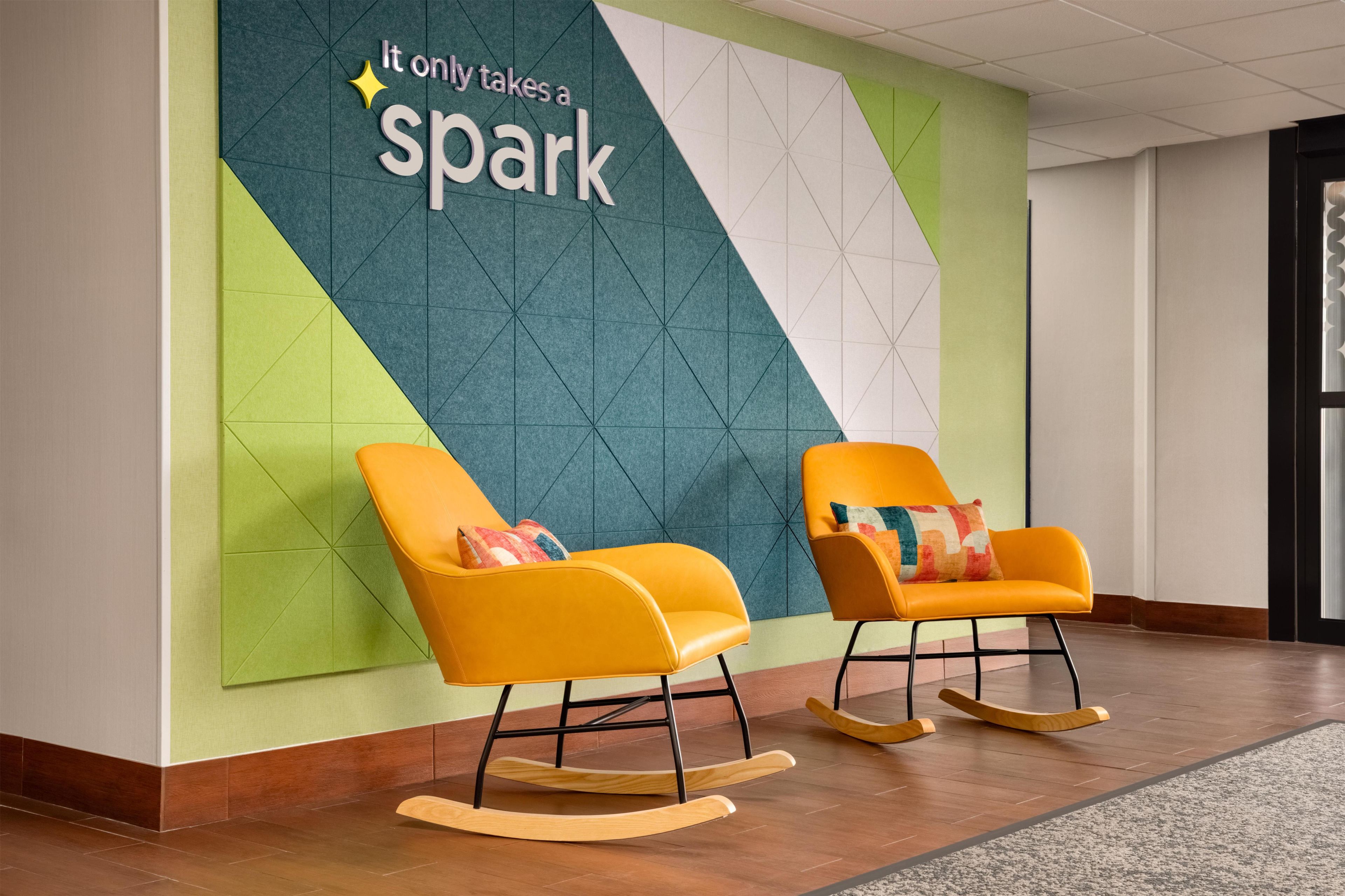 A modern lobby with two rocking chairs, a geometric wall design, and a sign that reads 'It only takes a spark'.