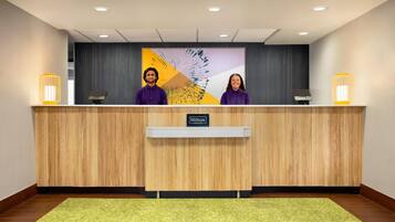 Un mostrador de recepción con dos recepcionistas, un mostrador de madera, una alfombra verde y una pared con un cuadro.