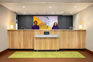 Vastaanottotiski, jossa on kaksi vastaanottovirkailijaa, puinen vastaanottotiski, vihreä matto ja seinä, jossa on taulu.