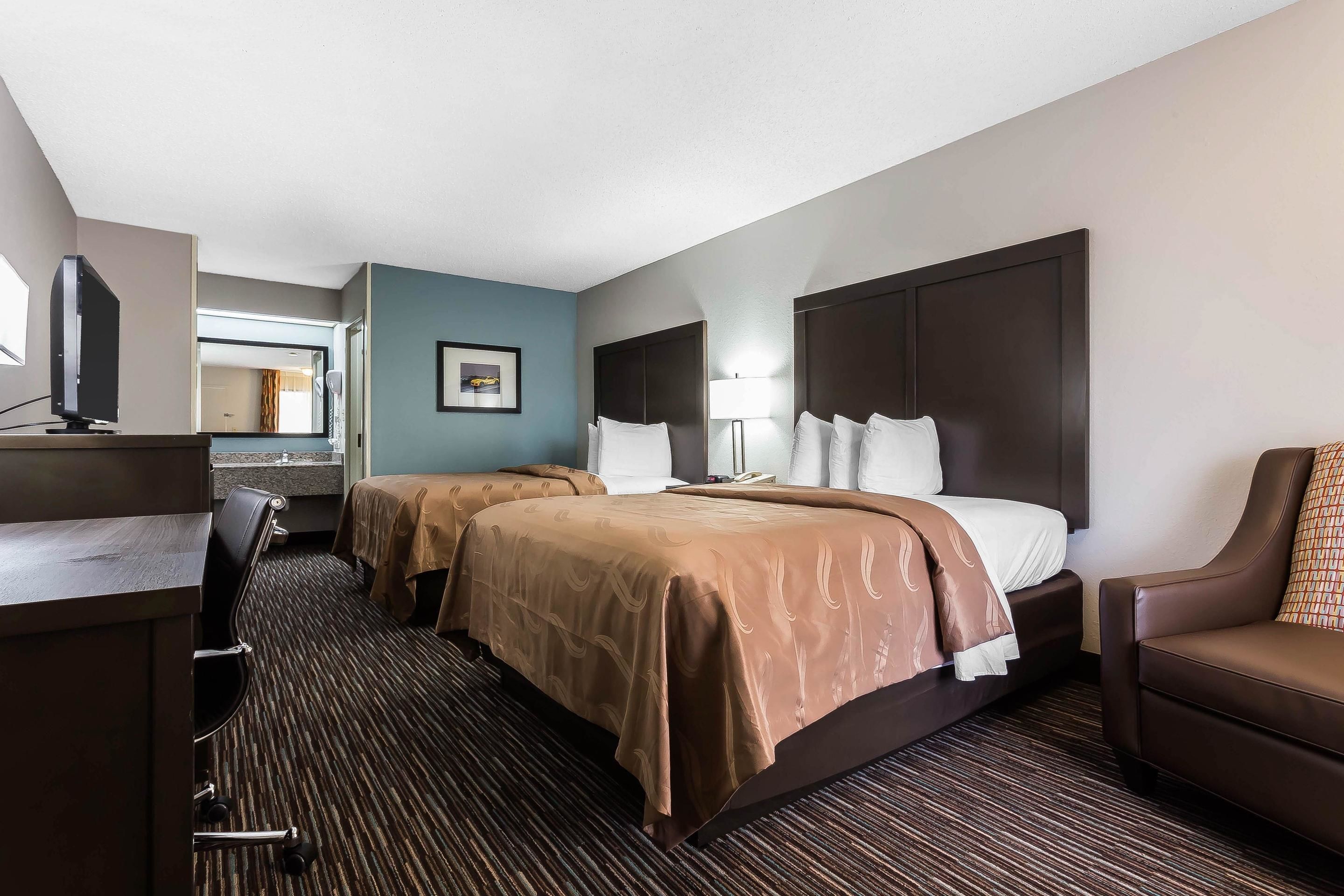 A hotel room with two beds, a desk, a chair, and a TV.