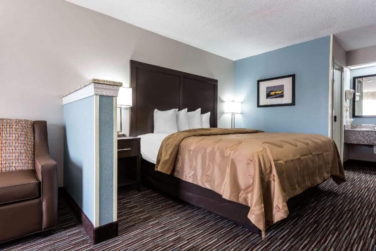 Habitación de hotel con cama, una silla, mesita de noche, lámpara, un cuadro en la pared y un baño con espejo y lavabo.