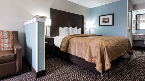 Une chambre d’hôtel avec un lit, une chaise, une table de chevet, une lampe, un tableau encadré au mur et une salle de bain visible à travers une porte ouverte.