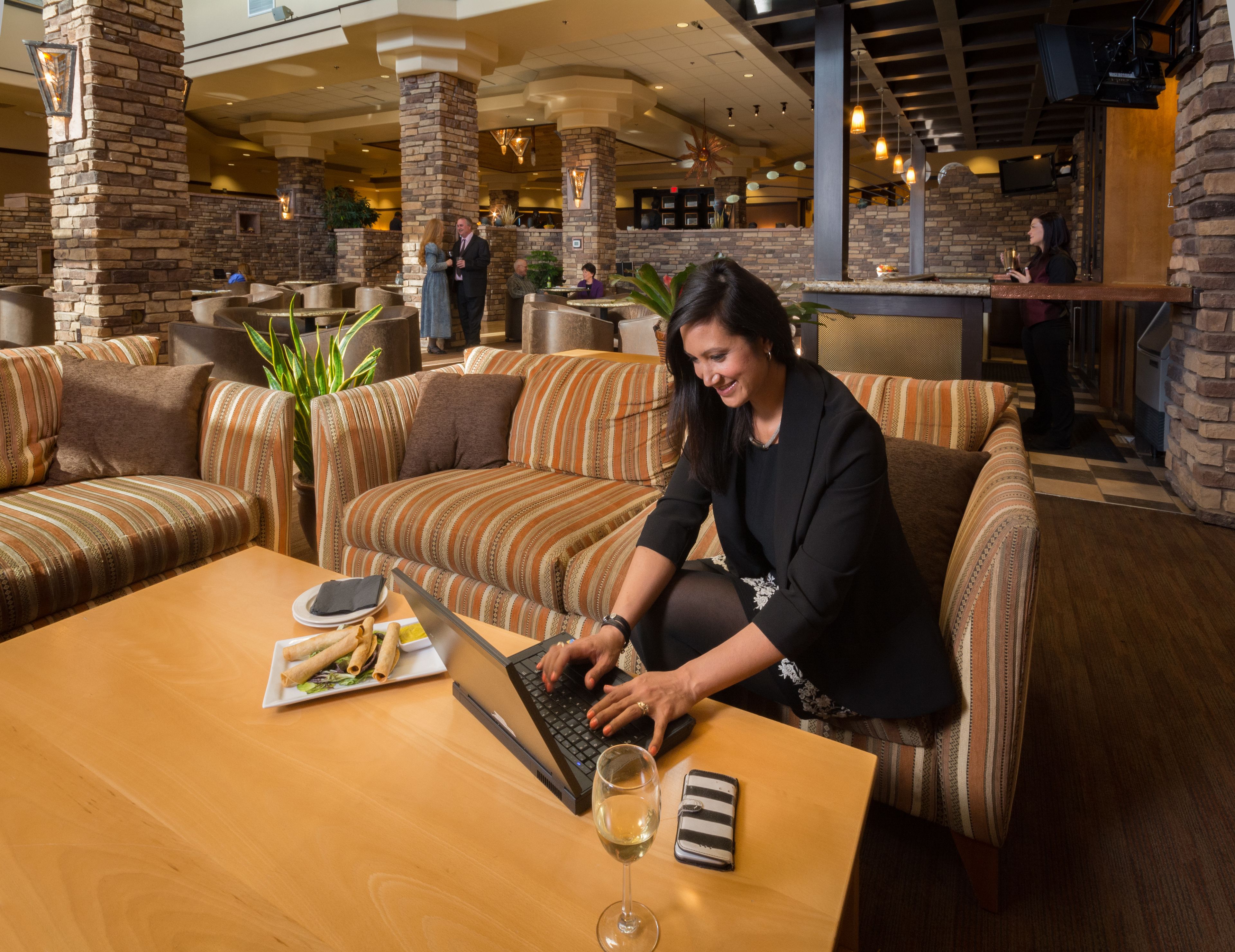 A woman sitting on a striped sofa at a hotel lobby with a laptop, a glass of wine, and a plate of food.