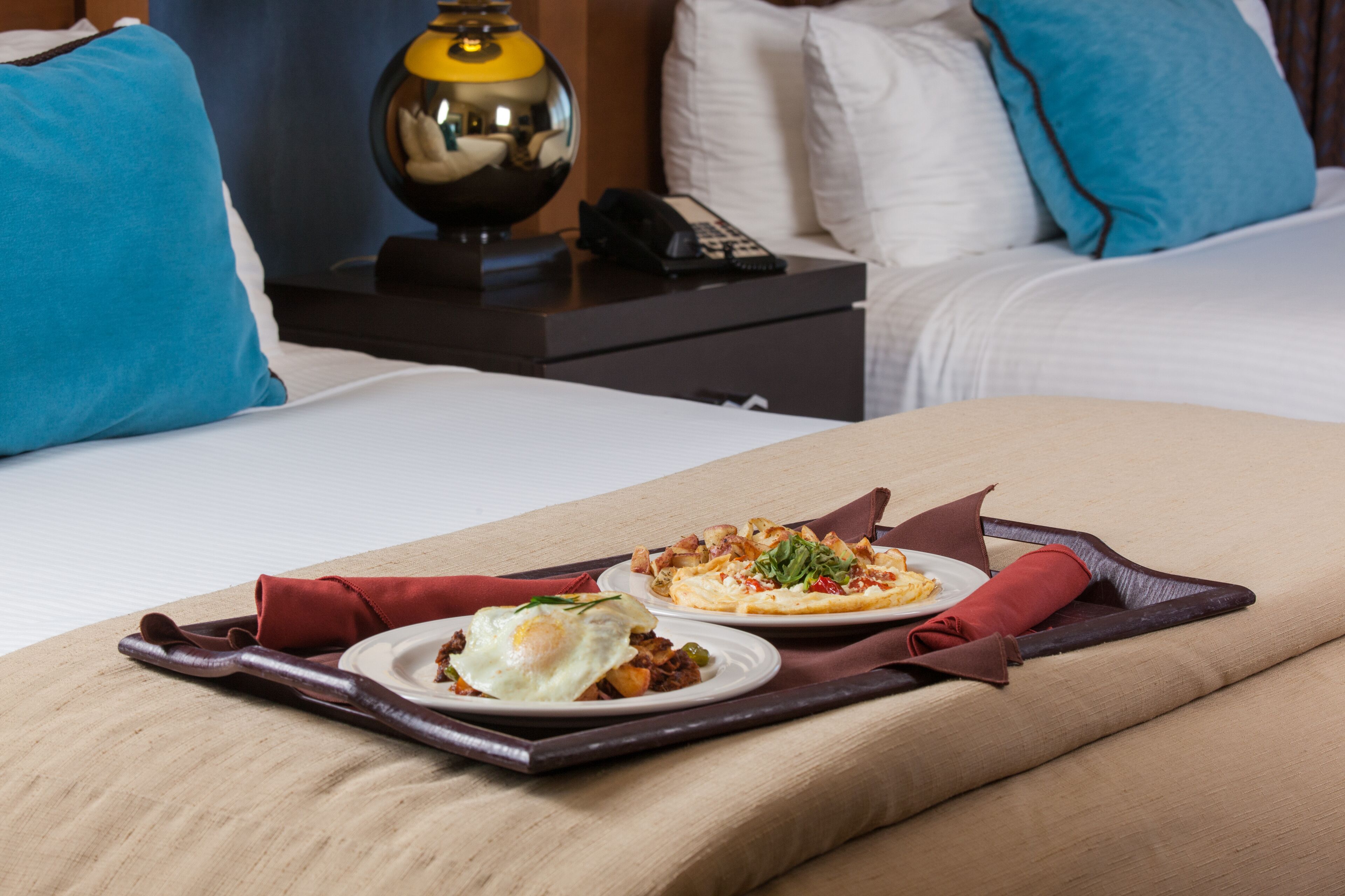 Een hotelkamer met een bed, een nachtkastje met een lamp, en twee borden ontbijt op een dienblad.