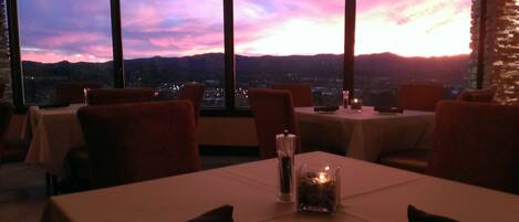 A restaurant with tables set for dining, large windows offering a view of the cityscape and mountains during sunset.