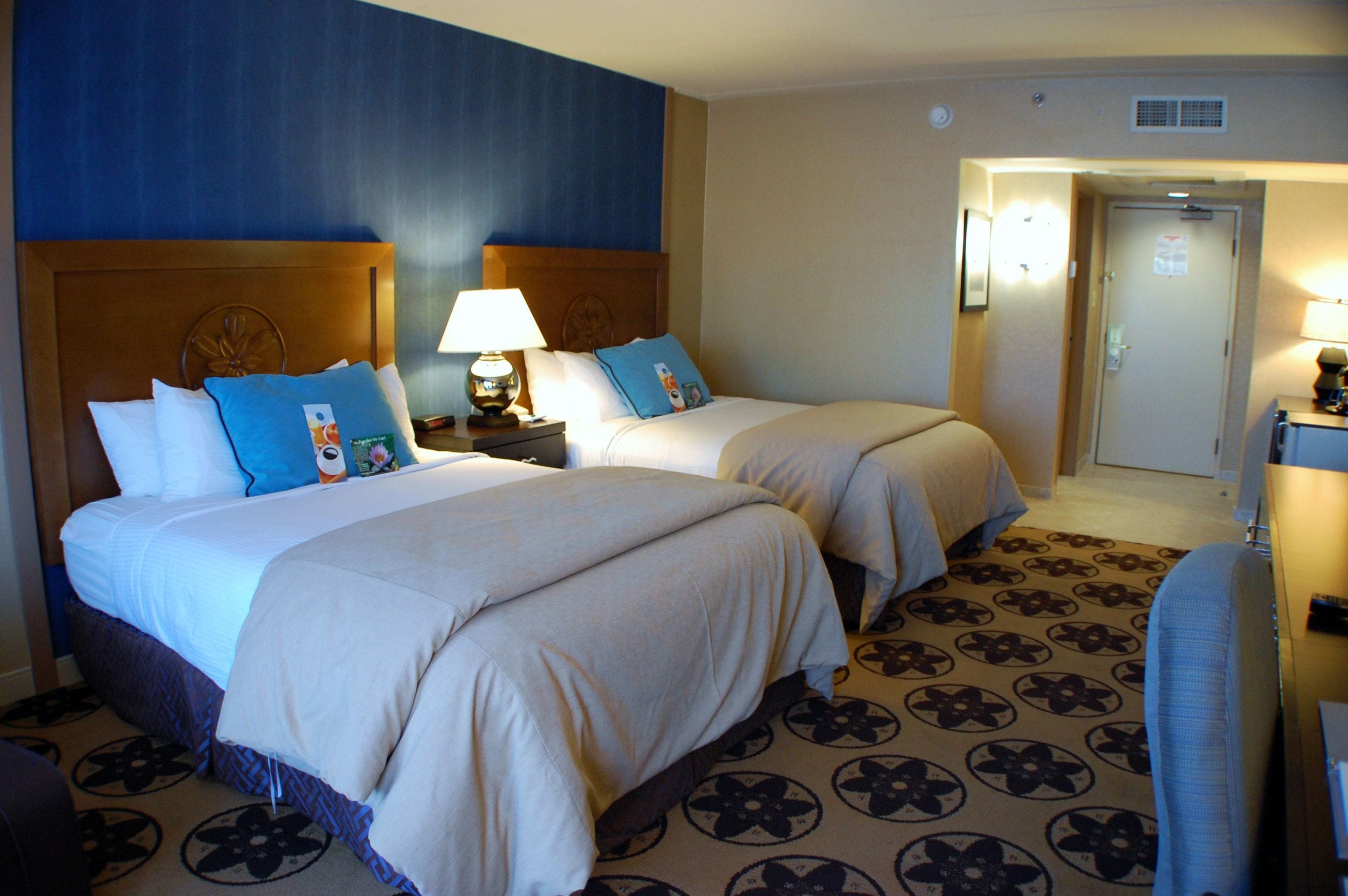 Quarto de hotel com duas camas, um tapete estampado e uma mesa de cabeceira com abajur.