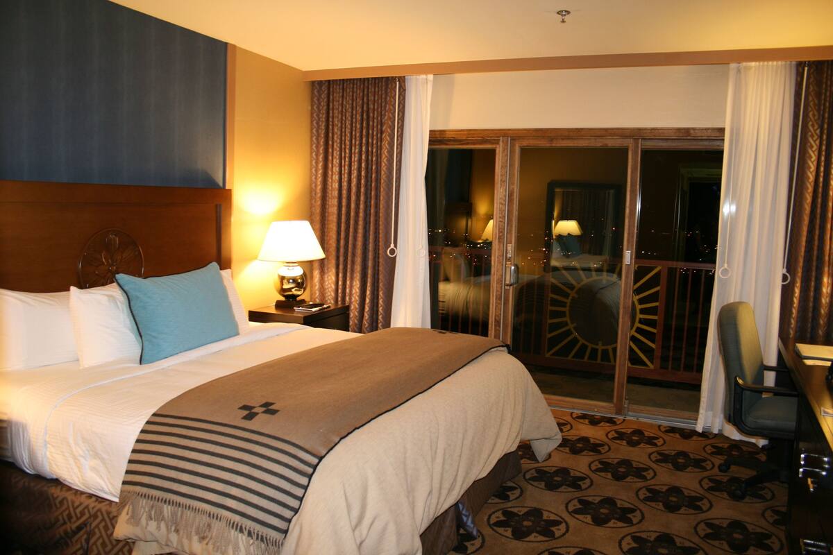 A hotel room with a large bed, a sitting area with a chair, a desk, and a view of the city through a glass door.