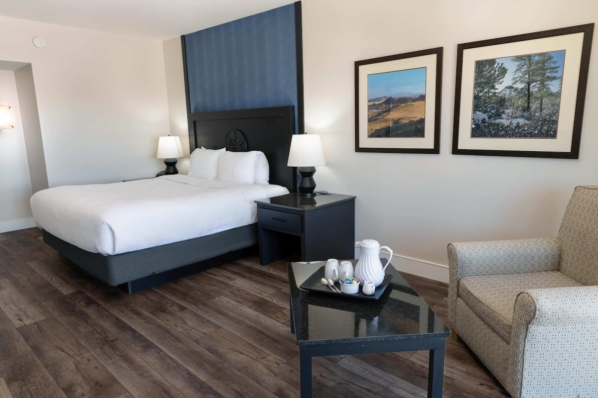 A hotel room with a large bed, two bedside tables with lamps, a coffee table with a tray of mugs, a sofa, and two framed pictures on the wall.